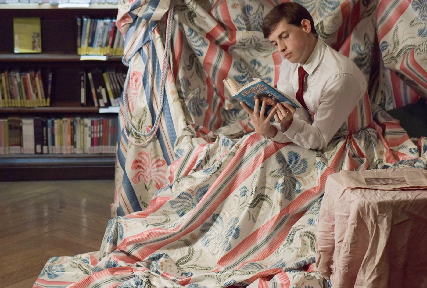 Moritz Gottschalk trägt ein weißes Hemd mit roter Krawatte und sitzt, ein Buch über Elizabeth Taylor lesend, eingehüllt in dicken bunten Stoff im Lesesaal der Bibliothek.