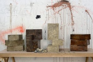 Die Farbfotografie zeigt vier Betonskulpturen in unterschiedlichen Grautönen von Rouven Dürr, die aus jeweils zwei bis drei Quadern zusammengesetzt sind. Sie stehen auf einem einfachen, aus Holzplatte und Böcken bestehenden Tisch. Die Atelierwand im Hintergrund weist Farbspritzer auf, das in zarten Versalien geschriebene Wort „schauen“ ist erkennbar.