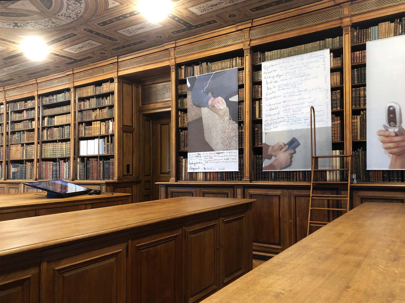 Ausstellungsansicht in der Universitätsbibliothek: an den hölzernen Regalen hängen Ausstellungsobjekte vor den Büchern