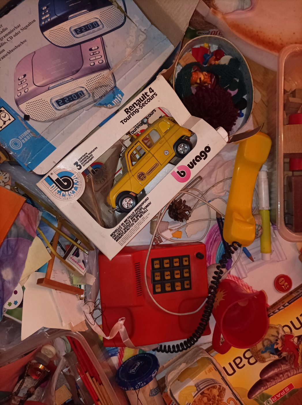 Die Fotografie zeigt ein rotes Spielzeugtelefon, ein gelbes Spielzeugauto in der Verpackung mit der Aufschrift "Renault 4", die Verpackung eines Radioweckers auf dem ein Blauer und rosafarbiger Radiowecker abgebildet ist, rund herum liegen Stifte und Papier und anderer Kram neben einer Packung Schokobananen und einem roter Trinkbecher