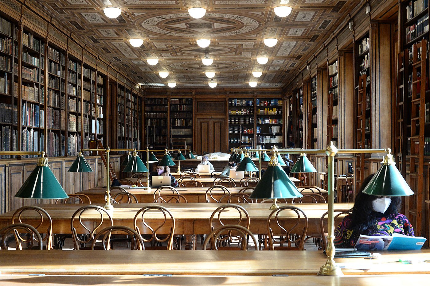 Historischer, von Theophil Hansen gestalteter Lesesaal der Akademie der bildenden Künste Wien. Leser_innen mit Laptops und FFP2-Masken sitzen an den von der Sonne beschienen Tischen.