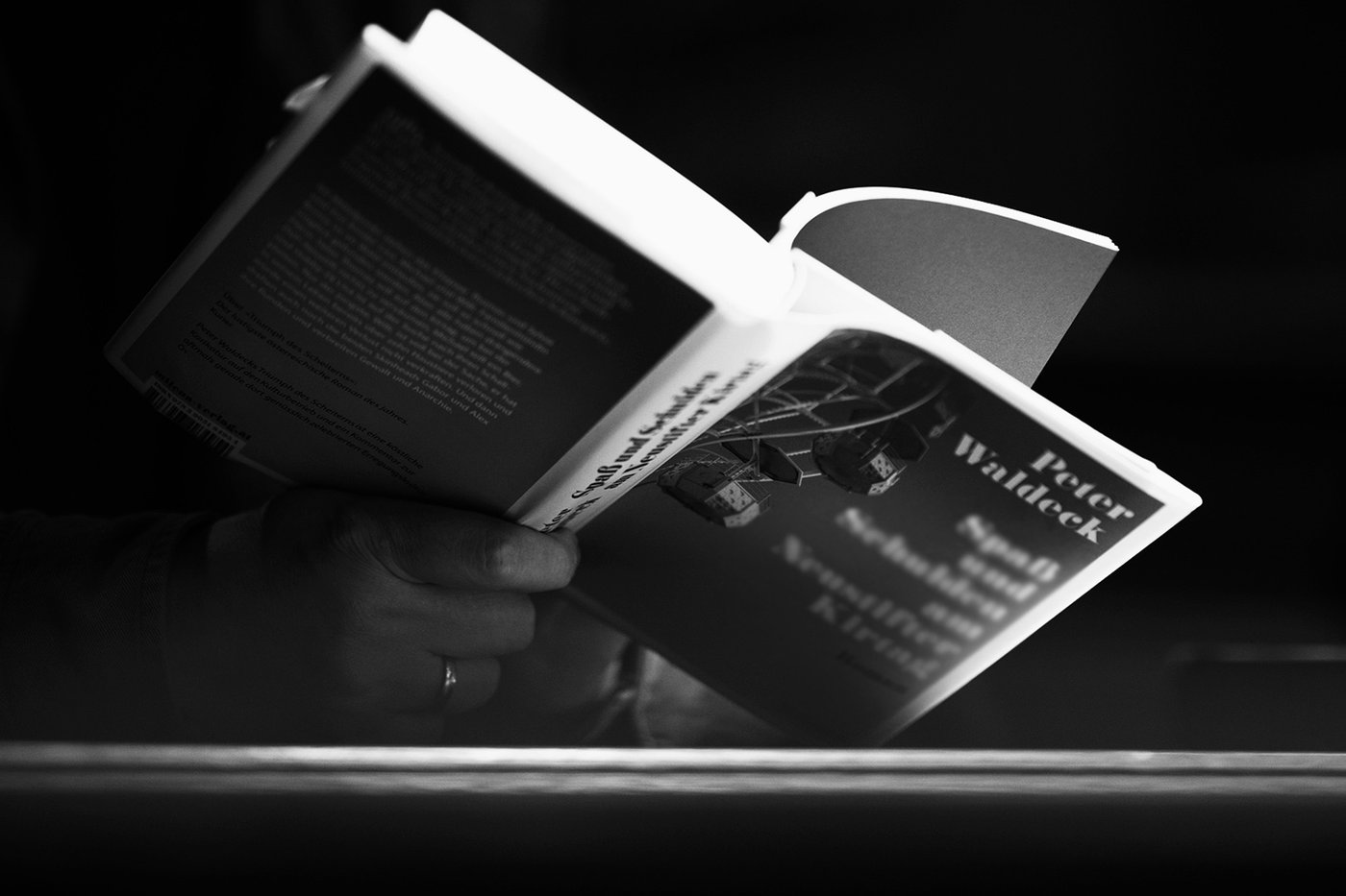 Die Schwarzweiß-Fotografie im Querformat zeigt eine Hand, die ein aufgeschlagenes Buch hält. Zu erkennen ist der Name des Autors: Peter Waldeck.
