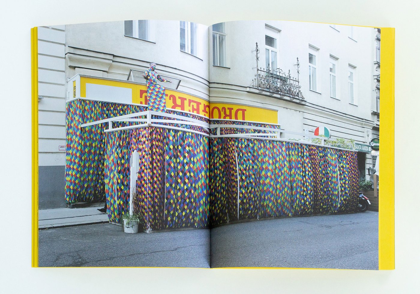 Die Farbfotografie im Querformat zeigt eine Doppelseite aus dem Katalog „Kulturdrogerie“ (2025) mit einer skulpturalen Arbeit von Thomas Hörl. Bunte Vorhänge verhüllten 2020 die Schaufenster des Artist-run Space Kulturdrogerie sowie ein Metallgestell davor. Darüber thronte ein Harlekin.