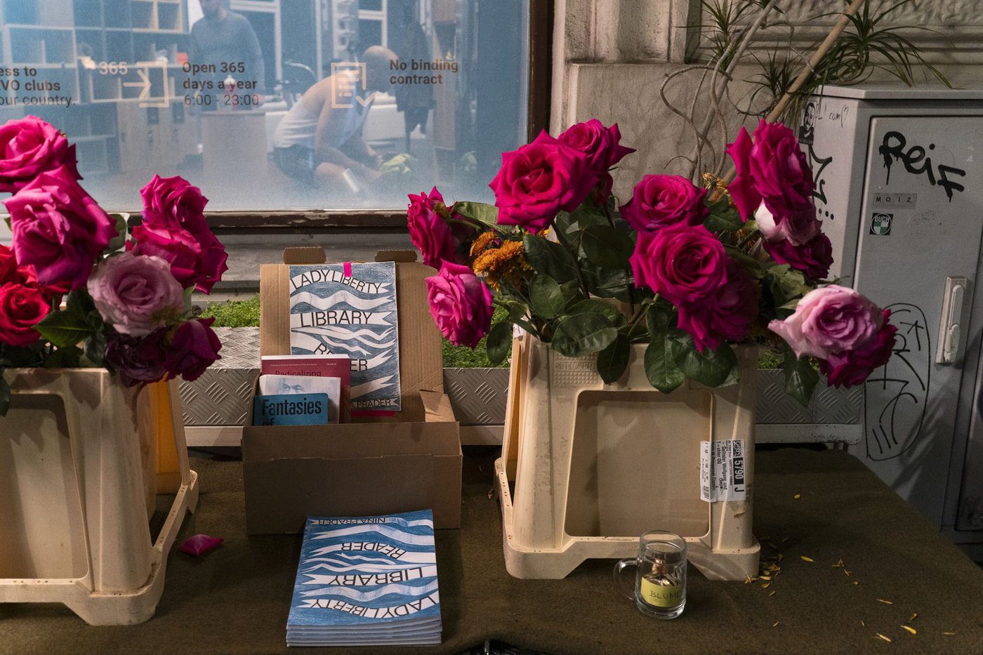 Die Farbfotografie im Querformat zeigt Nina Praders „Lady Liberty Library Reader“ in einer Bücherkiste zwischen zwei Plastikcontainers mit Rosen in voller Blüte. Im Hintergrund ist das Fenster eines Lokals mit Aufschriften wie „Open 365“ oder „No binding contract“ erkennbar.