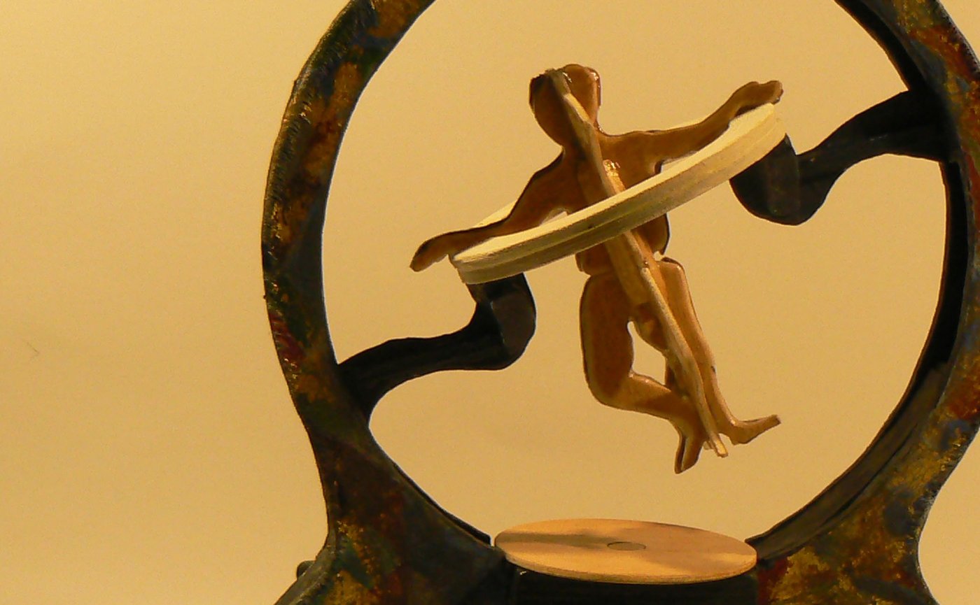 Die Farbfotografie im Querformat zeigt die Kleinskulptur „Sophrosynergon“ für die Lese-Performance „Sophrosyne“. Eine schräg gestellte Figur aus Buchenholz mit drehbaren Bögen erinnert an einen aus dem Lot geratenen „Vitruvianischen Menschen“.
