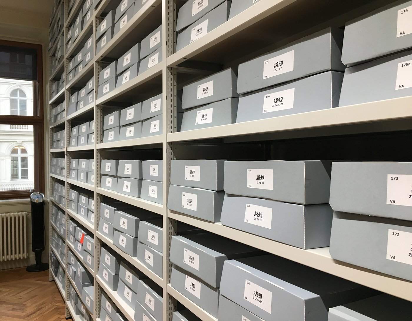 Innenansicht des Universitätsarchivs, zu sehen ist ein Regal vollgefüllt mit grauen Kartons, alle gleich groß und beschriftet mit bedruckten Etiketten.