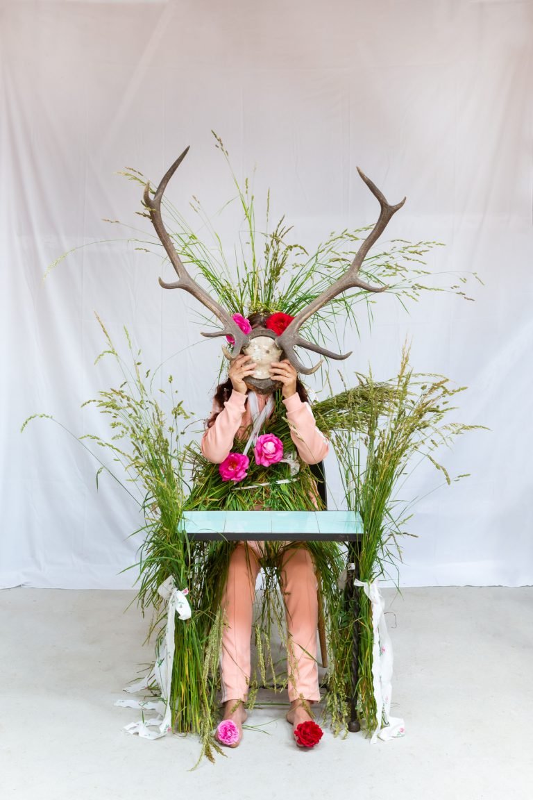 Frau mit Blumen und Gräsern und einem Hirschgeweih vor dem Gesicht, an einem Schreibtisch