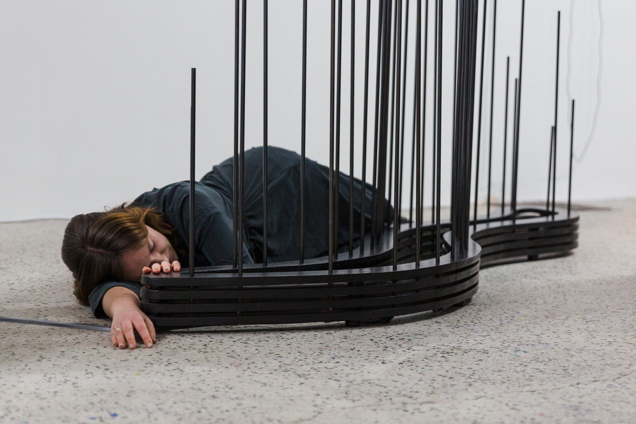 Frau in grauem Kleid, die am Boden liegt hinter einer Installation eines schwarzen Objekts mit vielen dünnen senkrechten Streben