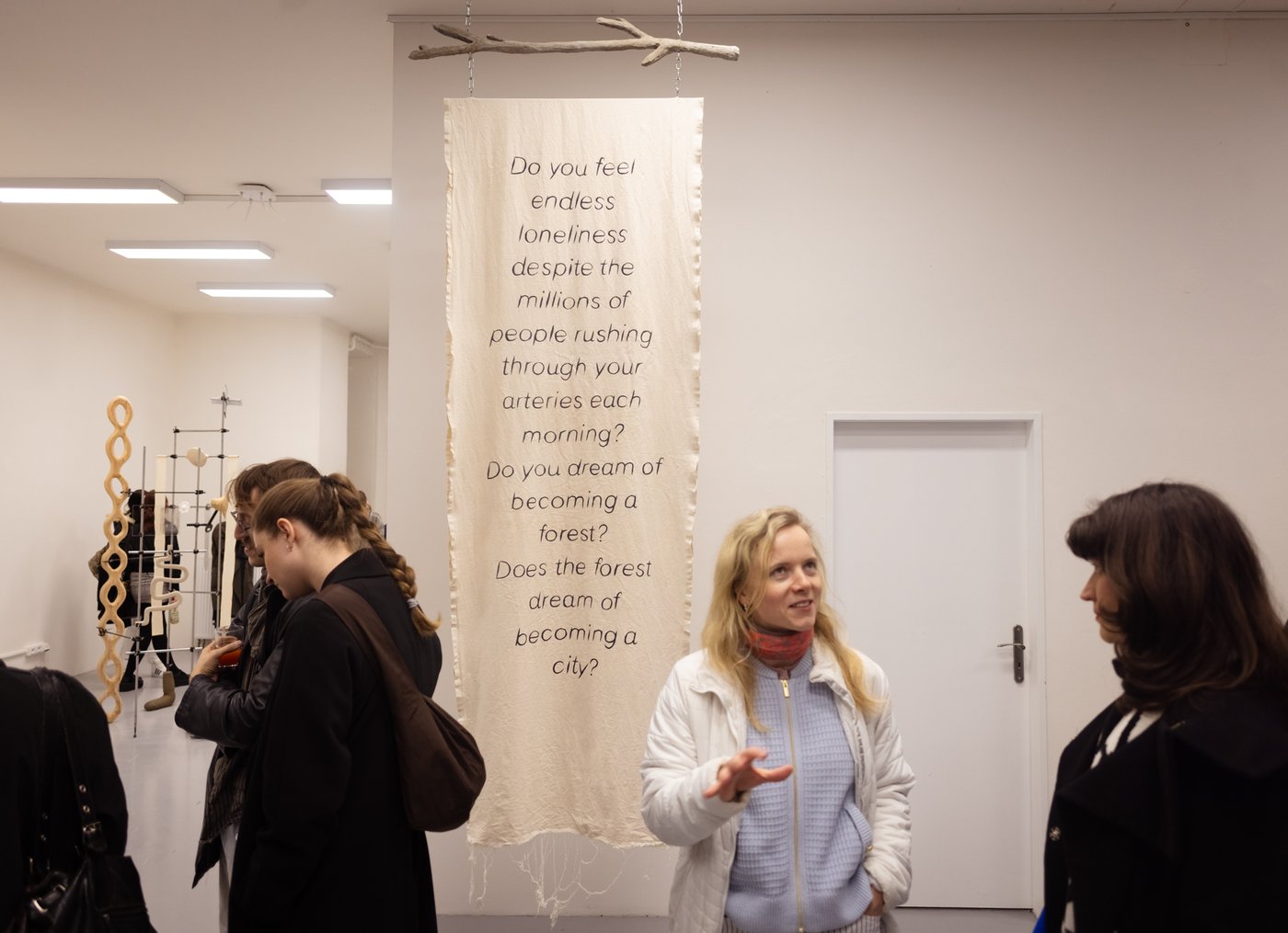 heller Stoff mit schwarzer Schrift hängt im Ausstellungsraum, herum Personen die das Kunstwerk betrachten