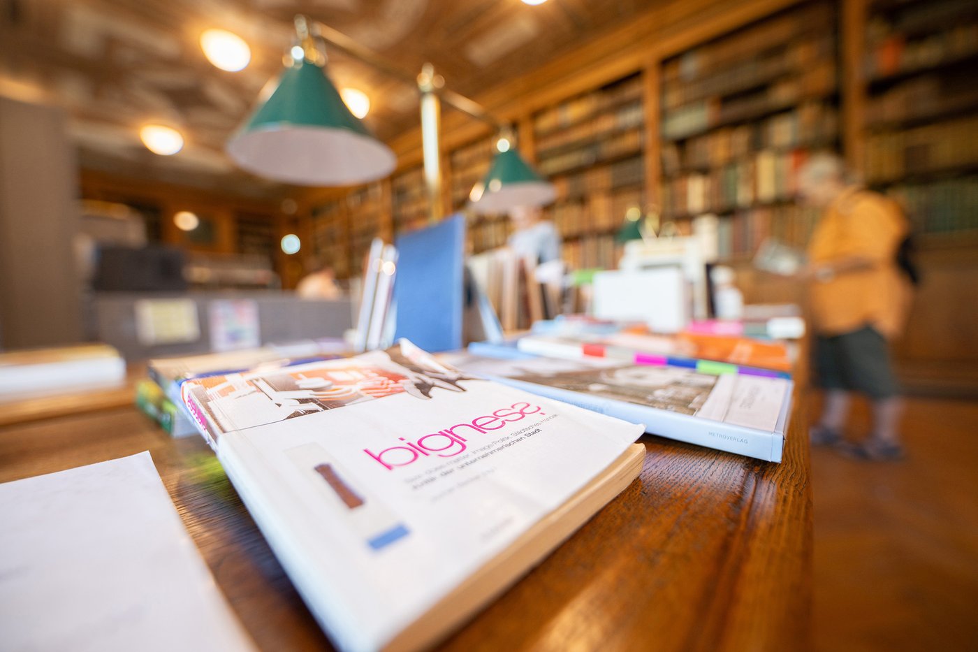 Die Farbfotografie im Querformat zeigt den von Theophil Hansen konzipierten Lesesaal mit historischem Buchbestand. Auf einem Originaltisch mit Leselampen im Vordergrund ist ein buntes Angebot von teils liegenden, teils stehenden Büchern präsentiert.