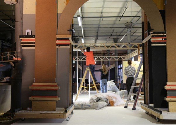 Im Vordergrund sieht man den Ausschnitt eines Tores aus Holz. im Hintergrund arbeiten drei Studierende am Aufbau einer Holzkonstruktion.