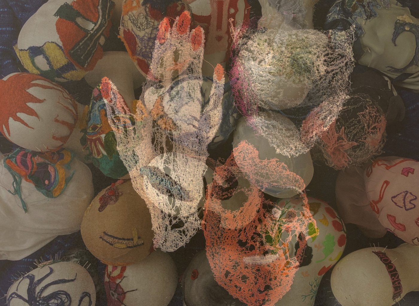 Das Bild zeigt eine Ansammlung von vielen runden Styroporköpfen, die dicht aneinander liegen. Die Köpfe sind mit farbenfrohen, textilen Stickereien versehen – darunter abstrakte Formen, Muster und Gesichter. Darüber liegt eine halbtransparente zweite Ebene aus maschinengestickten Motiven. In dieser überlagernden Schicht sind zwei filigrane, netzartige Gesichter sowie eine ebenfalls gestickte Hand zu erkennen. Die Stickereien bestehen aus dünnen, Faden-ähnlichen Linien, die ein luftiges, durchscheinendes Erscheinungsbild erzeugen. Insgesamt entsteht ein vielschichtiges, textil geprägtes Bild, das sowohl dichte Farben als auch transparente Strukturen zeigt.
