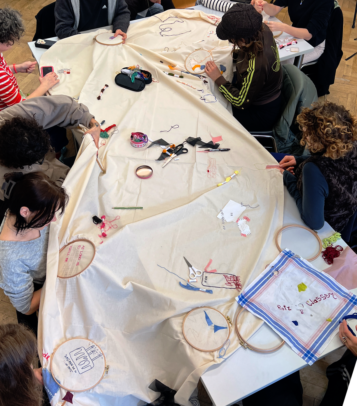Studierende sitzen um einen Tisch und besticken gemeinsam ein großes Stück Stoff.