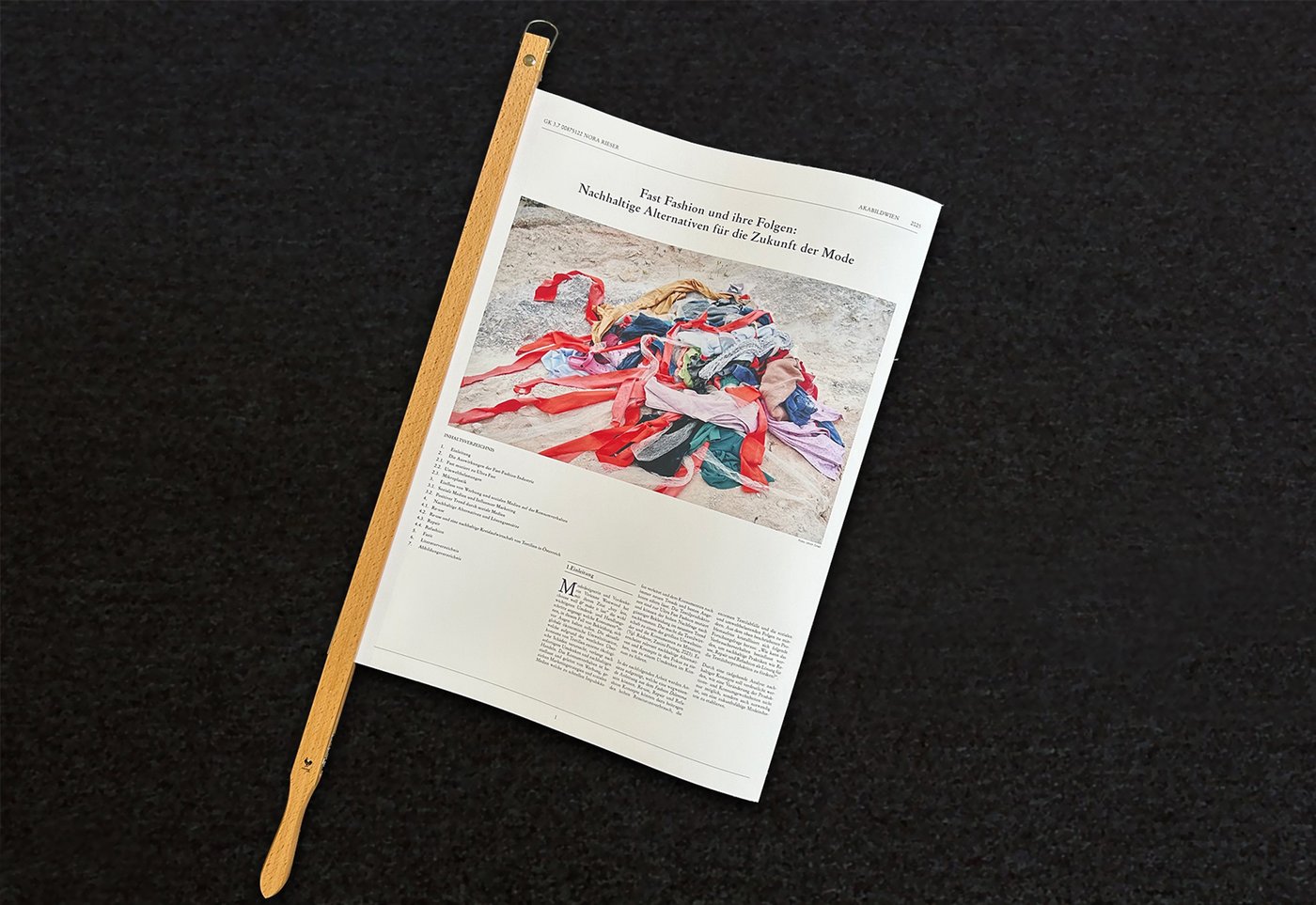 Nora Rieser, Seminararbeit „Fast Fashion“ in Zeitungsform, 2025Zeitung an der linken Seite in einem hölzernen Zeitungshalter​ eingespannt. Oben auf der Titelseite befindet sich eine große Fotografie eines bunten Textilhaufens auf sandigem Untergrund. Darüber steht der Titel „Fast Fashion und ihre Folgen: Nachhaltige Alternativen für die Zukunft der Mode“. Darunter folgen Inhaltsverzeichnis und zweispaltiger Text.