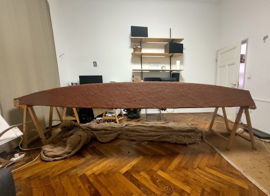 Das Bild zeigt den Prototyp eines kleinen Bootes, das aus natürlichen Materialien gefertigt wurde. Der braun beschichtete Bootsrumpf liegt verkehrt auf Holzböcken zur Bearbeitung auf.