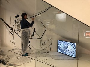 Installationsansicht mit Künstler der gerade dabei ist die Wand mit schwarzem Stift zu bemalen, in der rechten Ecke steht ein Bildschirm mit gezeichneter Figur
