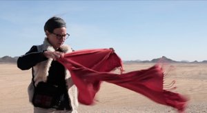 Weiblich gelesene Person steht in einer kargen Umgebung und hält einen roten Schal in den Wind