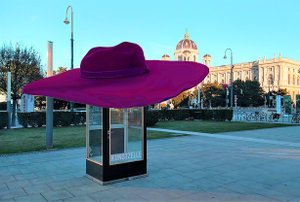 Eine Telefonzelle mit Aufdruck “Kunstzelle” steht am Vorplatz des Museumsquartiers. Sie trägt einen großen lila Hut, der ihr mit einem Fotobearbeitungsprogramm aufgesetzt wurde.