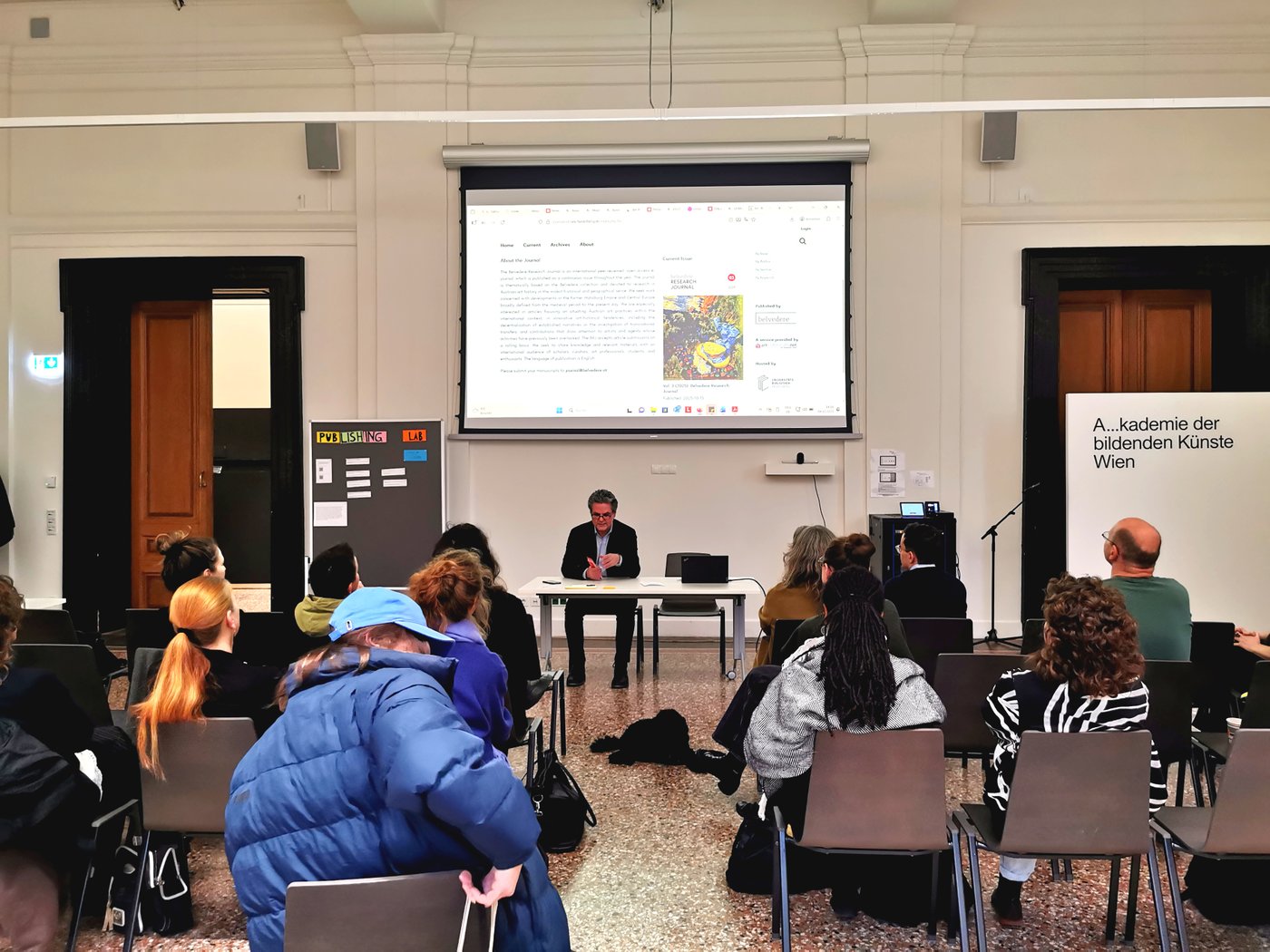 Eine Person sitzt hinter einem Tisch unter einer Projektion auf die Wand. Vor ihr sitzt Publikum.Neben dem Tisch steht ein Flip-Chart mit Zetteln und der Aufschrift "Publishing Lab".