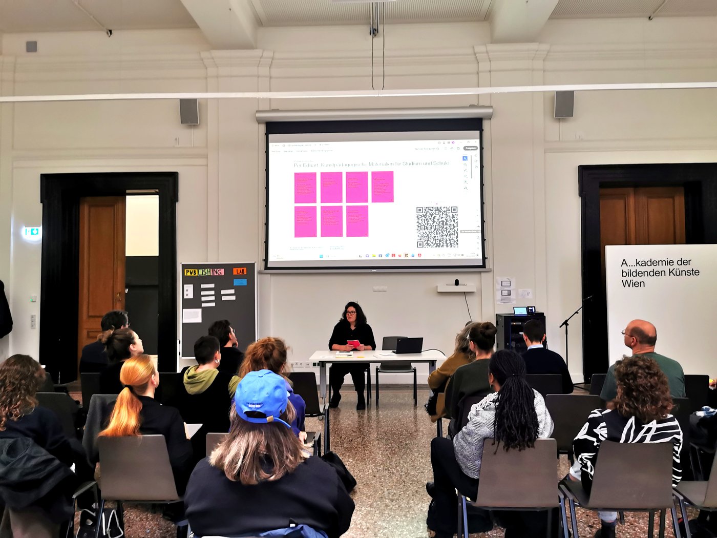 Eine Person sitzt hinter einem Tisch unter einer Projektion auf die Wand. Vor ihr sitzt Publikum.Neben dem Tisch steht ein Flip-Chart mit Zetteln und der Aufschrift "Publishing Lab".