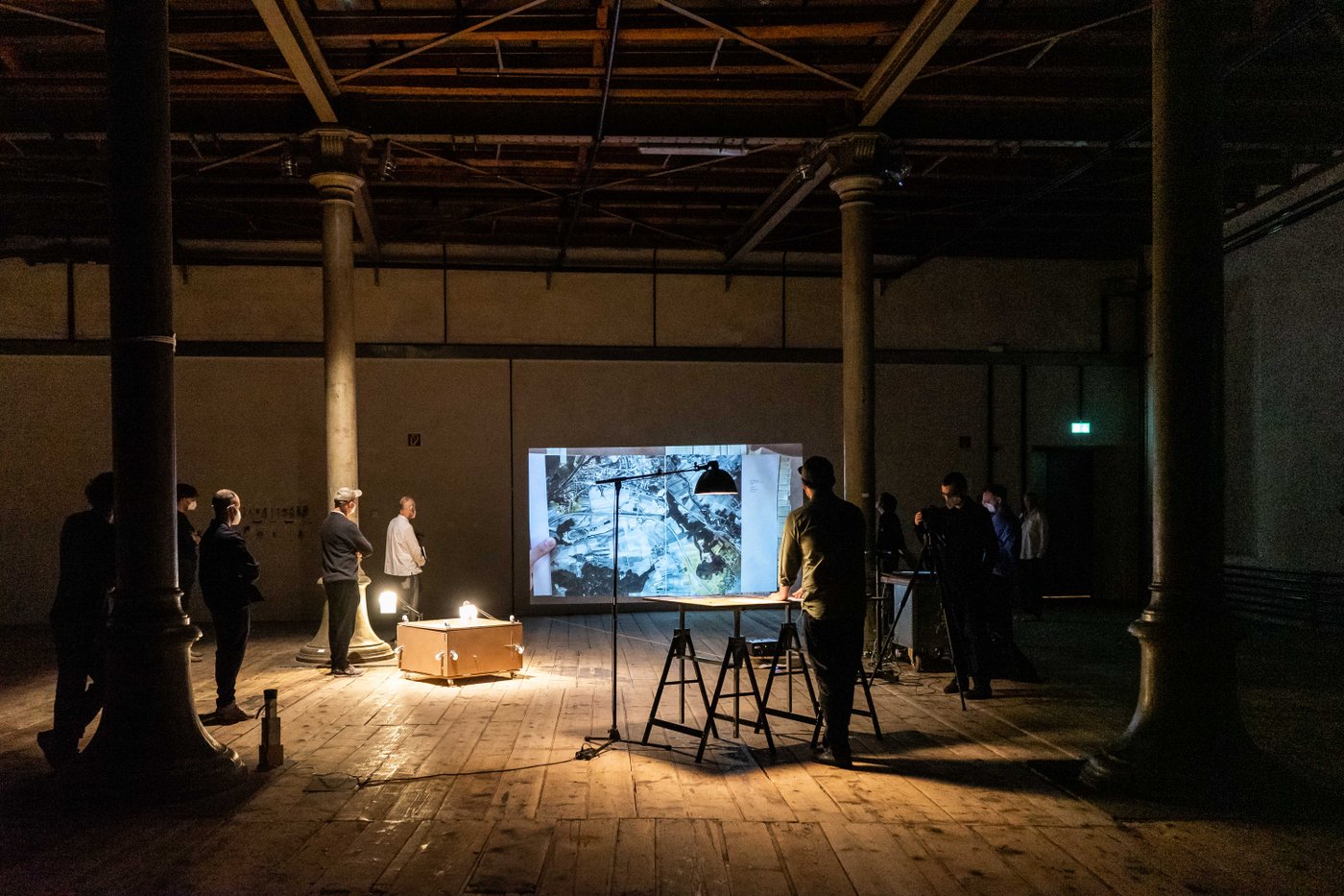 Diploma presentation in Semper Depot, a large, dark, open space with four pillars. In the foreground, one person stands at a table and talks about the project, which is projected onto the wall via beamer. To the left and right of the image are audience members.