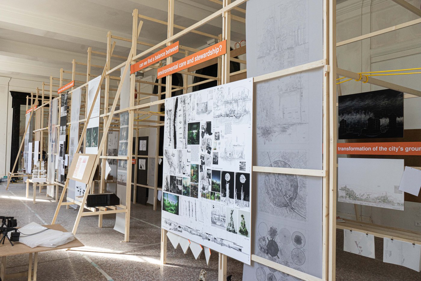 Exhibition view of the IKA's HITZE exhibition, various plans and drawings are mounted onto an almost room-high wooden scaffolding. An orange panel in the centre of the picture reads "can we find a balance between environmental care and stewardship?