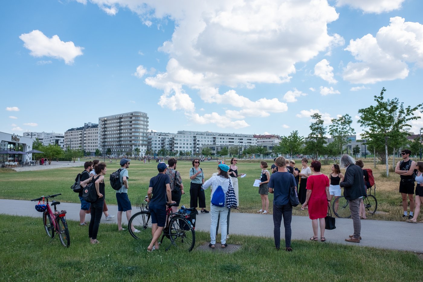A group of about 20 people are standing on a green lawn in Helmut-Zilk-Park in 1100 Vienna, in the “Sonnwendviertel” urban development area, listening to two people talking about the building in the background.