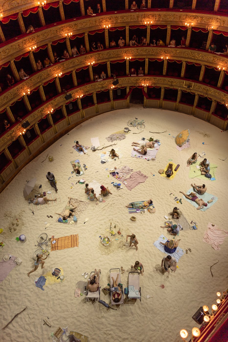 Interior of a theatre, audience looks down at people sitting in the sand.