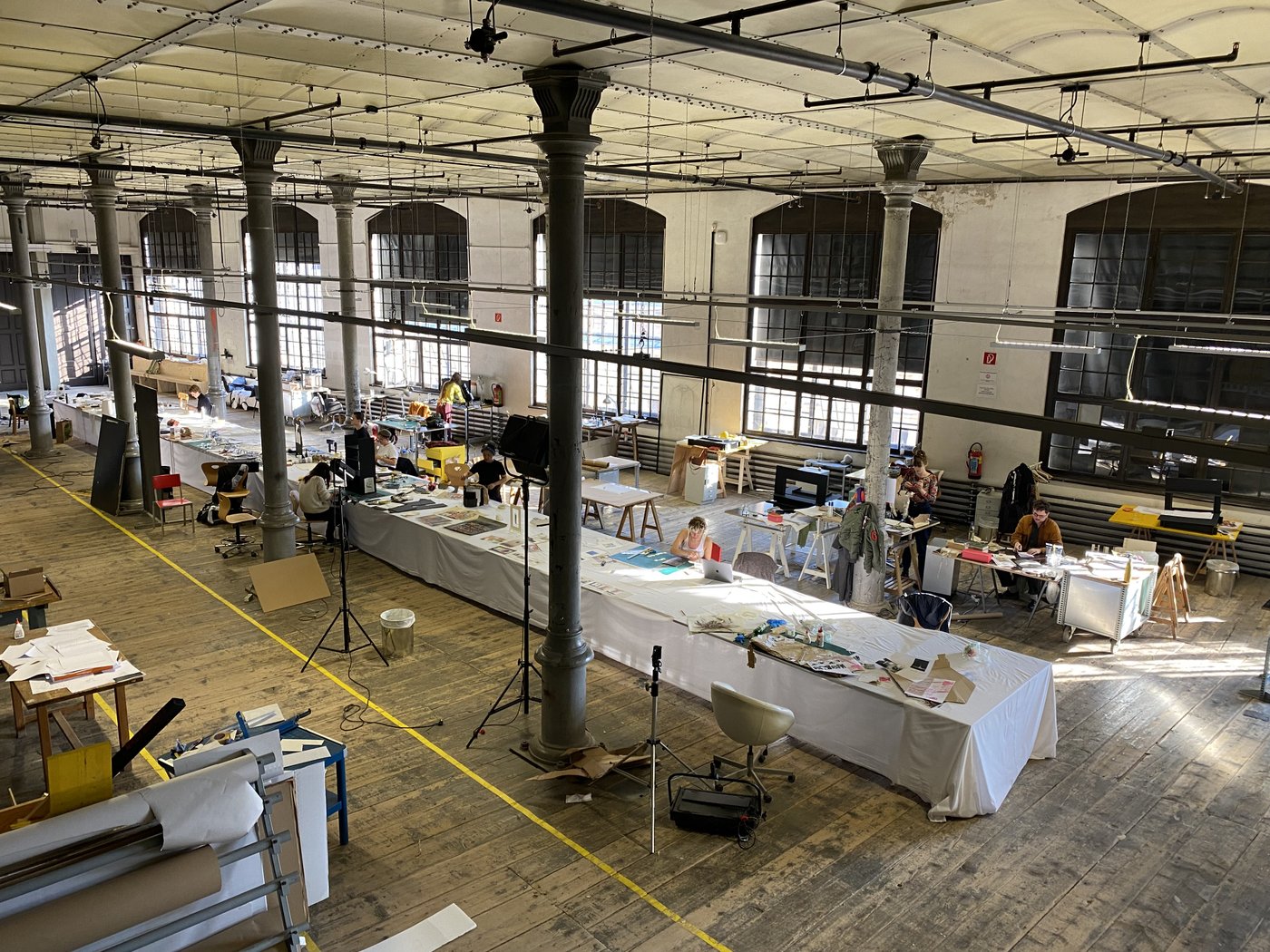 A long table stands in a huge room with rows of columns and 6 large windows. Students are working at this and other workstations, work material is lying and standing around everywhere.