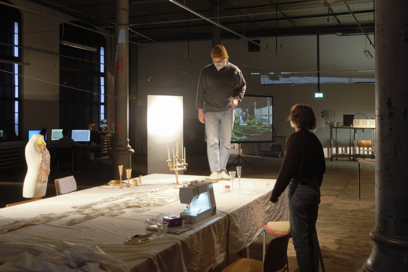 A student stands on a table covered with a cloth, on which a sewing machine, a candlestick, golden dishes and various other utensils lie. A female student is standing to the right. In the background you can see the studio room with a screen, a model and PC workstations.