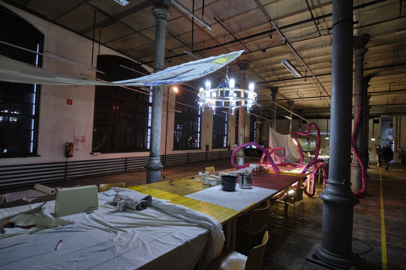 In the workroom of the stage design there is a long table with various working materials, a red paper snake, painted cloths stretched across the room above it and a chandelier.