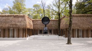 Photo of a building with thatched roof, behind it green trees, in the middle the building breaks backwards and gives the space for a large sharp figure with a large round circle as a head and hanging bosom, stairs lead to the elevation to the sculpture.