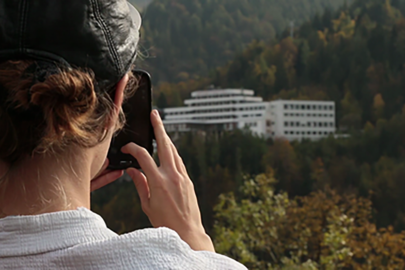Video still. In the foreground on the left, you can see the back of a person's head taking a picture with a black phone of the background, where a large white house with many windows can be seen in a hilly forest.