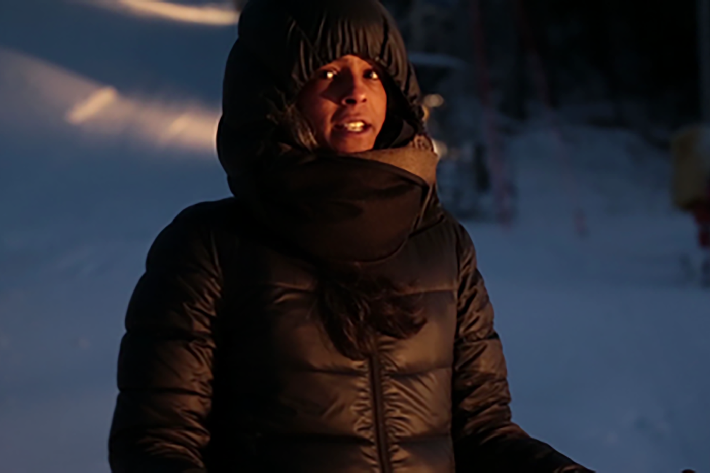 Video still. A person wearing a black down winter jacket and hood looks just past the camera with slightly open mouth and wide open eyes, in the background white landscape, probably snow-covered.