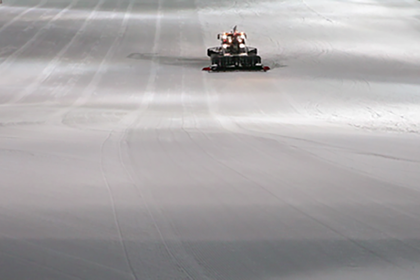 Video still. A large white area, a planned ski slope, further back a vehicle can be seen preparing the slope.