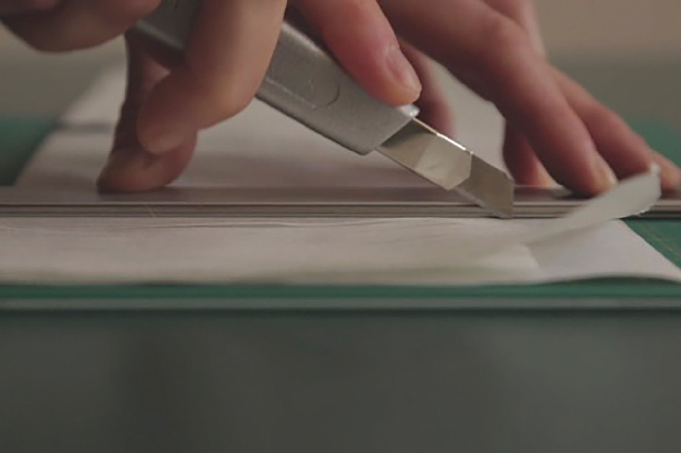 Hands can be seen using a silver paper knife and a ruler to cut through three superimposed pages of paper on a green cutting pad.
