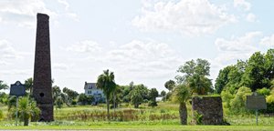 The image shows a historic site under a bright sky. A house is visible within a grassy field surrounded by palm trees, a tall brick chimney, a smaller brick ruin, and two historical information markers.