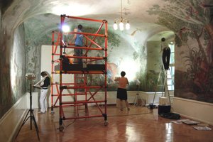 In a historic hall with ceiling and wall paintings, a rolling orange scaffold stands in the middle of the room; a student is working on it. In the background on the wall you can see three more female students, one of them is standing on a ladder in a window alcove.