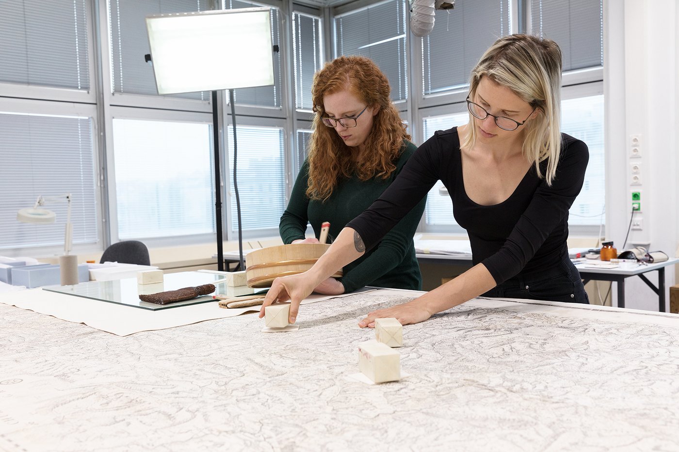 Two female students are working on a historical map lying on a large work table in the foreground of the picture. One student - standing behind the table on the left - is stirring the paste for the gluing in a container; the other - further to the right - is placing small cube-shaped weights on the glued crack areas.