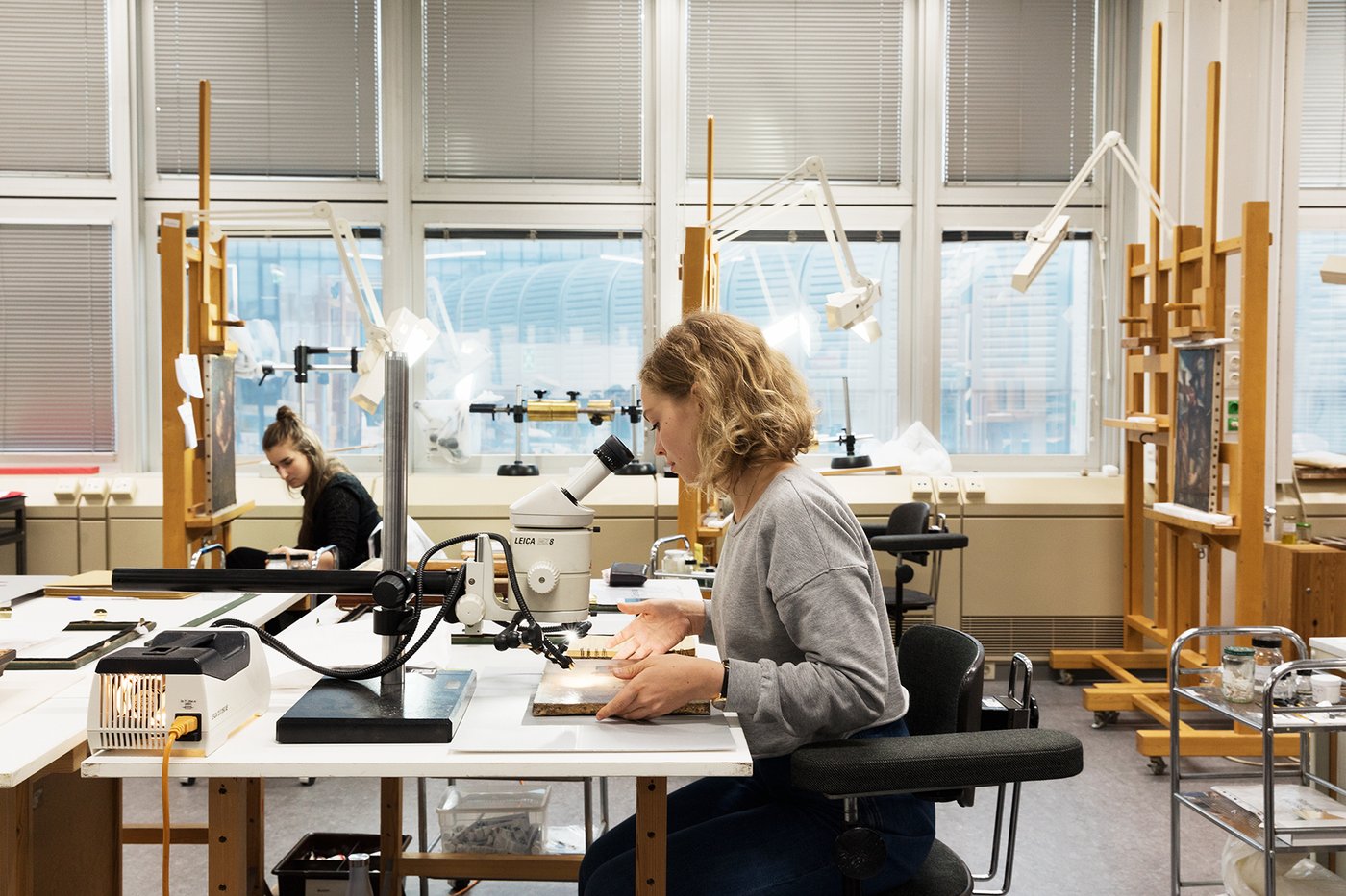 In the center foreground, a student is sitting at a work table; the photo shows her in profile from the left. In front of her is a small painting that she is sliding under a microscope. In the background on the left is a student in front of a painting on an easel, behind her the window front; on the right of the picture are more easels.