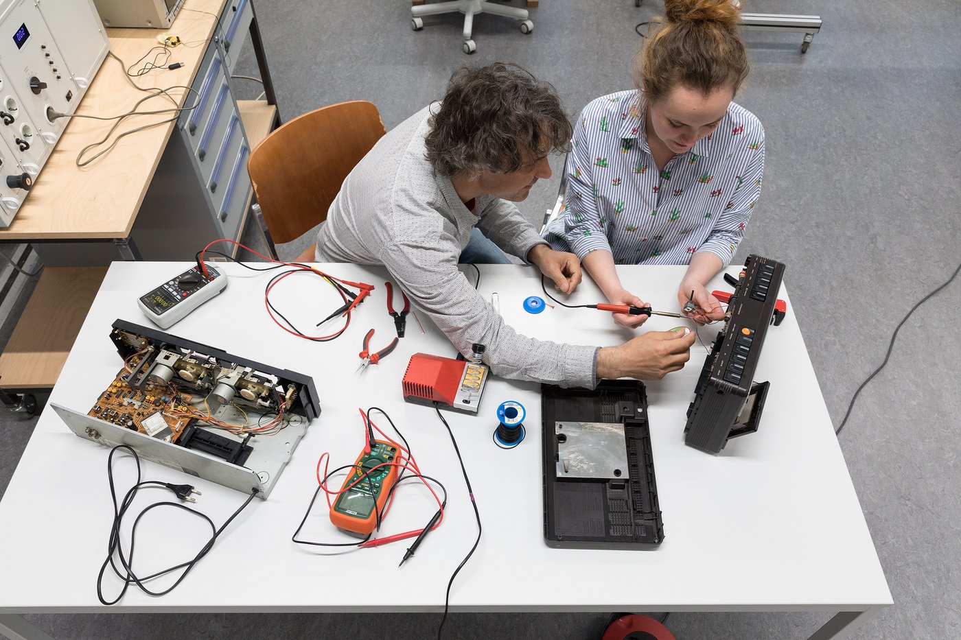 In the foreground of the image, a bird's eye view shows a bright work table on which a disassembled audio player and measuring devices are lying. A lecturer and a student sit side by side at this table and work together on the device.