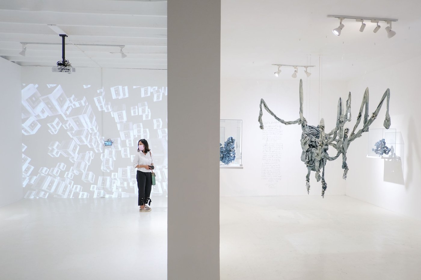 Exhibition view with blue-grey fabric bundles tied together to a large object and hanging from the ceiling, around small sotff bundles in glass showcases