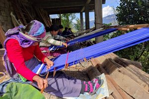 The photo shows 5 people sitting on the floor weaving different color fabrics on handlooms