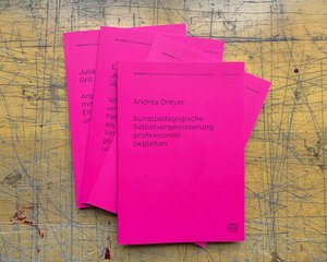 In the center of the picture four magenta colored booklets are stacked on a rugged wooden surface. The top booklet reads as follows: „Andrea Dreyer: Kunstpädagogische Selbstgewisserung professionell begleiten.“