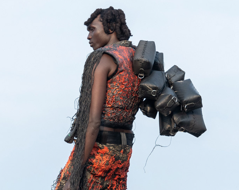 A person wearing an outfit made entirely from recycled materials—tire rubber, fishing nets, and orange fabric—stands in profile against a bright sky. On their back, they carry several black bags sewn from hoses.