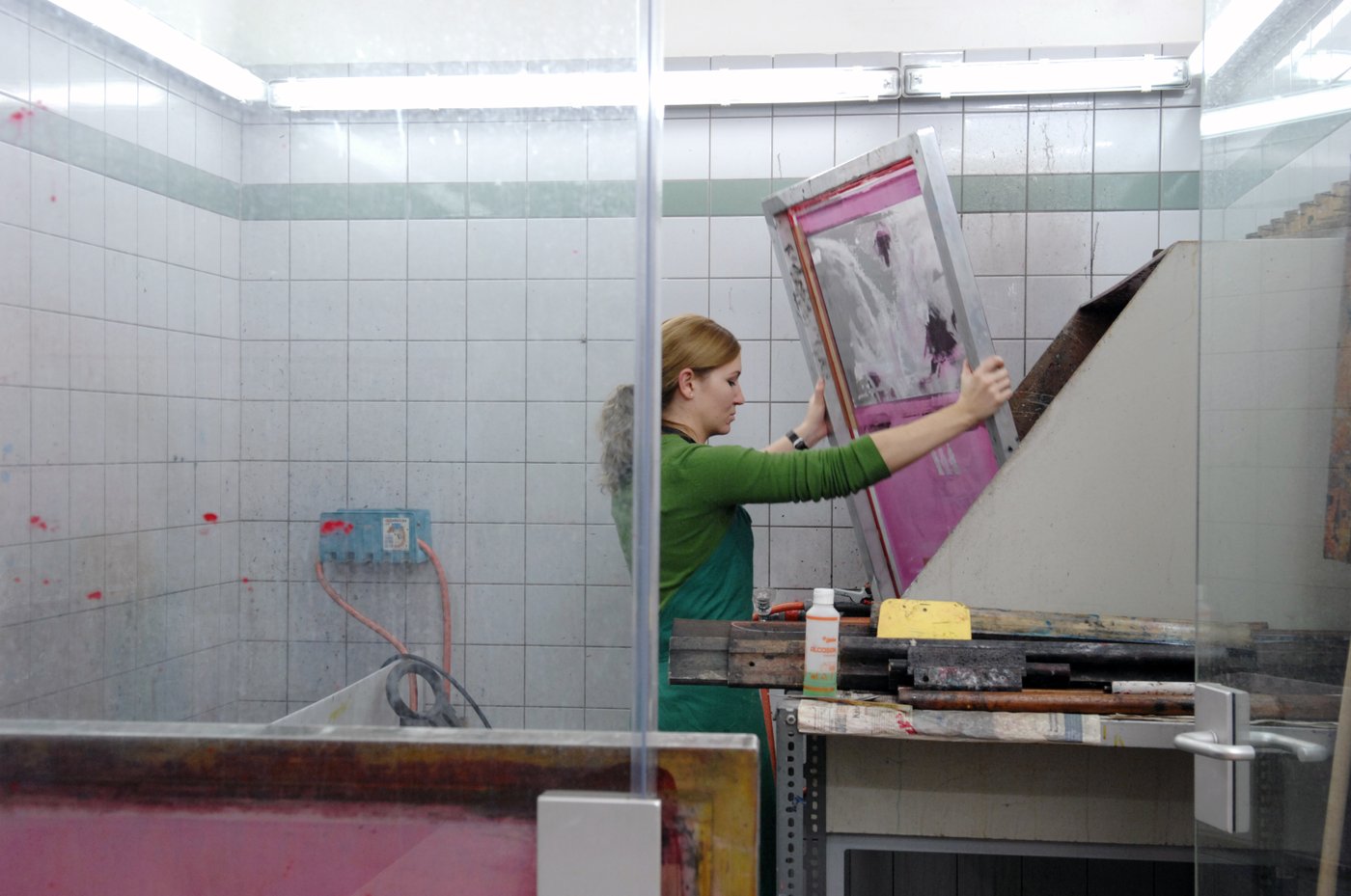A student is working in the screen printing workshop. She has a green apron on and is holding up the screen printing frame. The walls are tiled and partially glazed. There are all kinds of equipment lying around.