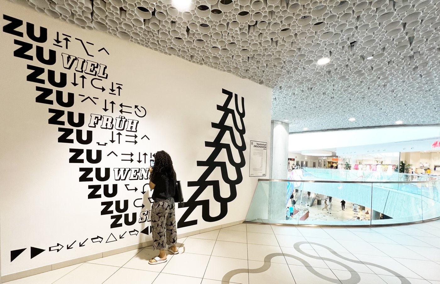 Cecilia Schaaf from hezelschaaf stands in front of the art wall and takes a picture of her artwork with her cell phone. To the right of the art wall is the mall and some customers are walking on the lower floor.