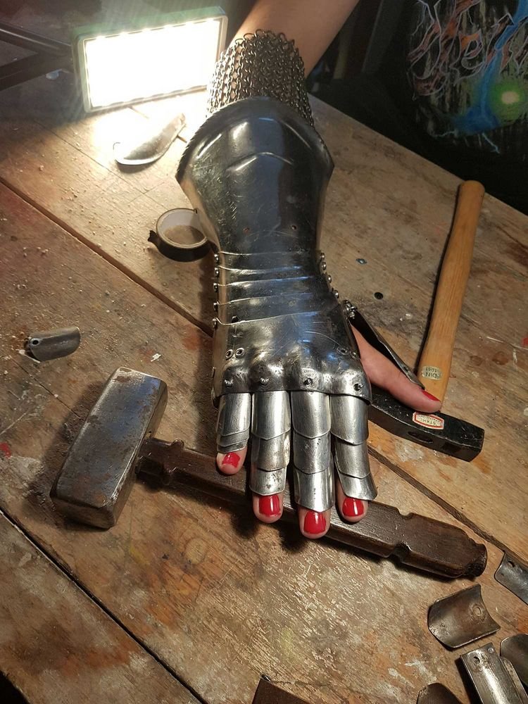 A hand in a gauntlet is placed on a table with different tools. The fingernails of the hand are painted in red.