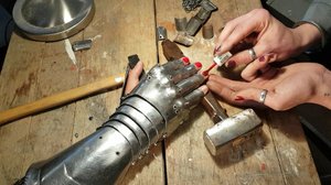 A hand in a gauntlet is placed on a table with different tools. The fingernails of the hand are painted in red.