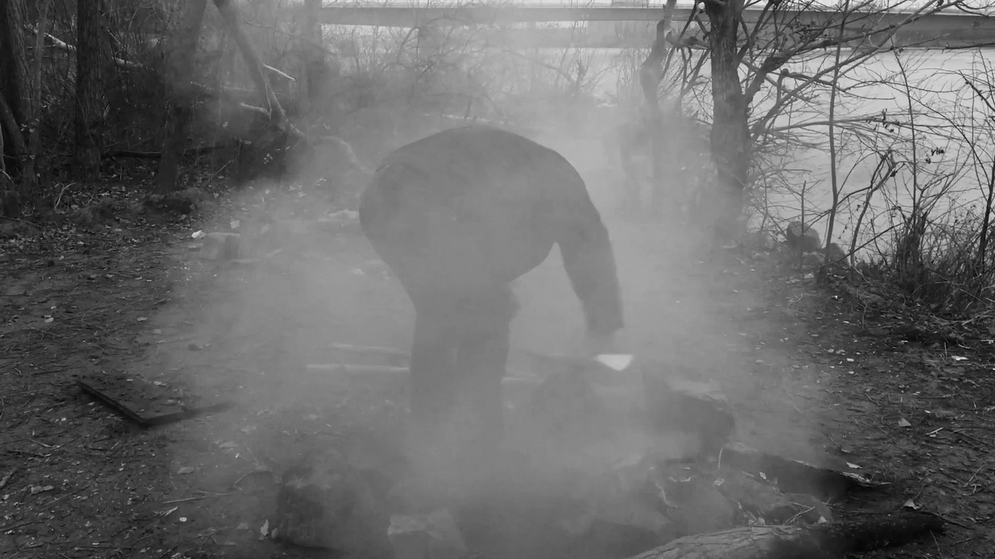 A part of the film “The Danube River”. A man is starting a little campfire by the river. The smoke is all around him. The smoke keeps going.